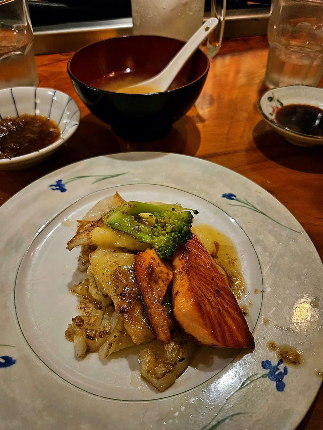 Grilled salmon fillet with broccoli, onion and buttery sauce on a blue-and-white plate, with miso soup and dipping sauces behind