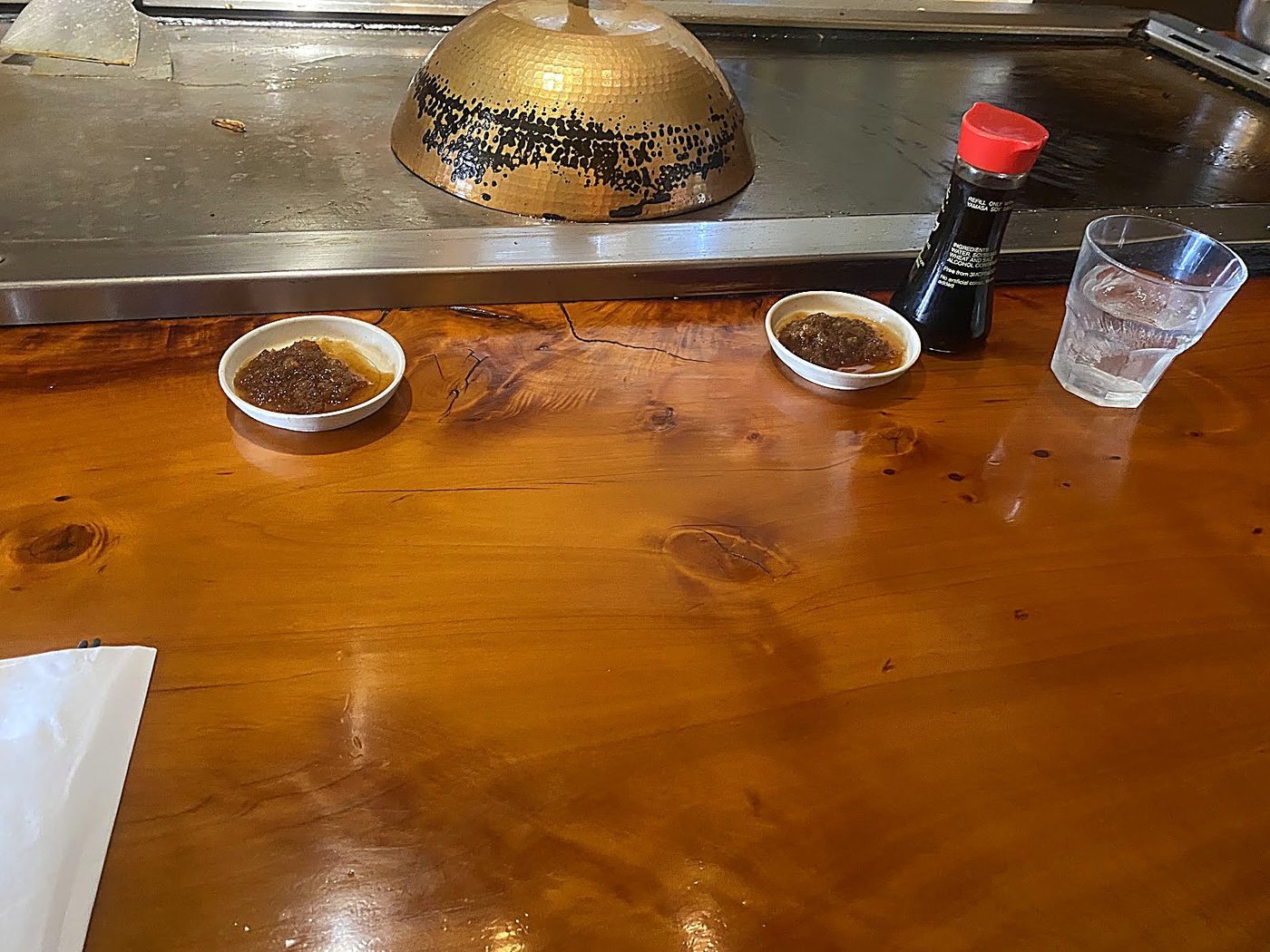 The polished kauri counter running alongside the teppanyaki griddle, with the brass dome resting on the hot plate and a soy sauce bottle set out