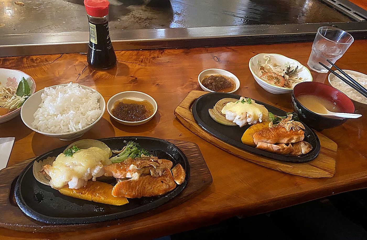 Two teppanyaki sets on cast-iron plates: prawns under melted yum yum sauce with grilled onion and pumpkin, salmon fillet, steamed rice, miso soup and green salad