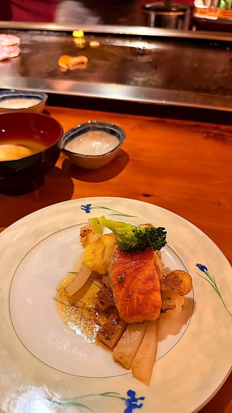 A plated teppanyaki main of salmon and grilled vegetables on a white plate, with miso soup and a small dish of sauce