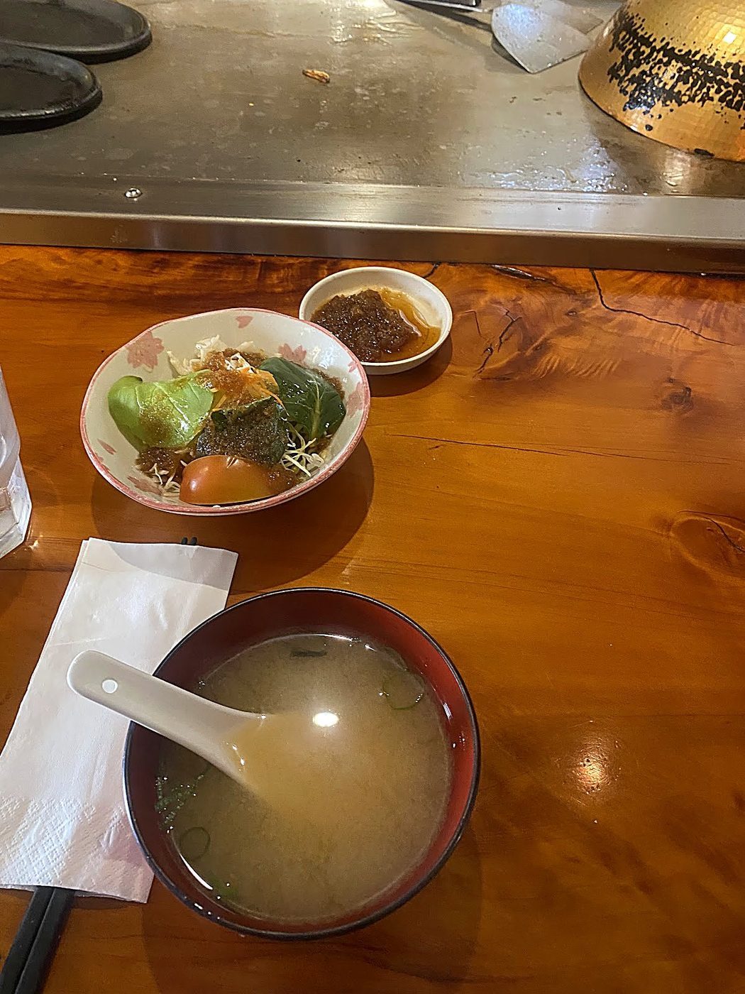 A small plate of grilled prawns and broccoli with a bowl of miso soup and a paper napkin on the counter