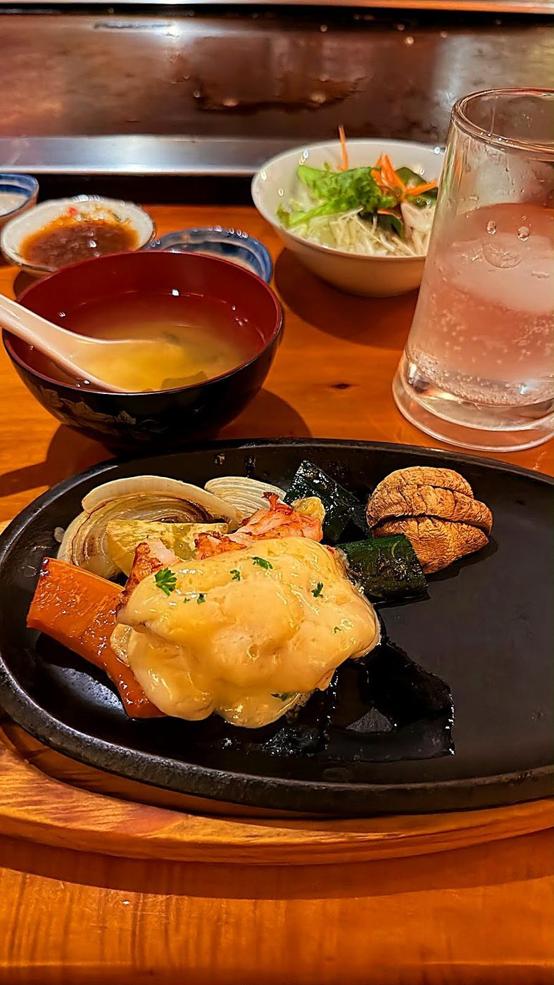 A teppanyaki plate of prawns and vegetables with miso soup and a cold drink on the warm-lit wooden counter